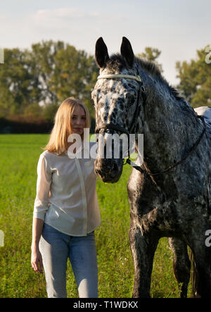 Jockey ragazza è delineare un cavallo su open manege. Bella ragazza jockey è in piedi vicino a un cavallo baciando e abbracciando il suo Foto Stock