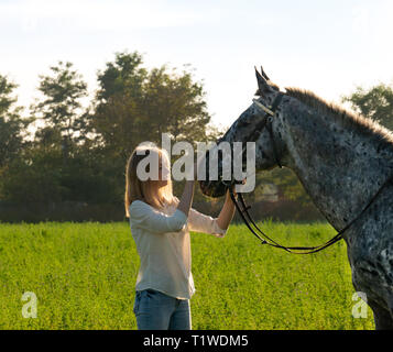 Jockey ragazza è delineare un cavallo su open manege. Bella ragazza jockey è in piedi vicino a un cavallo baciando e abbracciando il suo Foto Stock