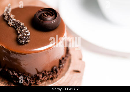 Una deliziosa torta al cioccolato con pezzetti di cioccolato risiede su un supporto di legno accanto a una tazza di bianco, che si erge su un bianco tavolo in legno Foto Stock
