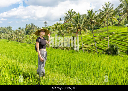 Alla moda rilassato caucasian turista femminile che indossa il piccolo zaino e tradizionali asiatici paddy hat passeggiando tra splendidi e verdi campi di riso e Foto Stock