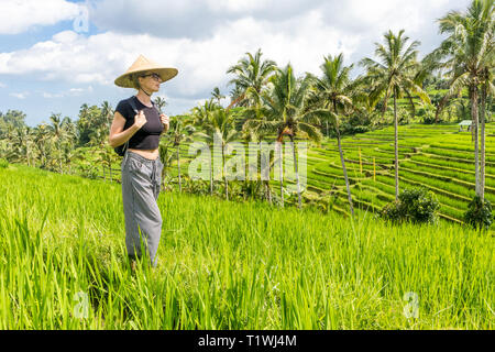 Alla moda rilassato caucasian turista femminile che indossa il piccolo zaino e tradizionali asiatici paddy hat passeggiando tra splendidi e verdi campi di riso e Foto Stock