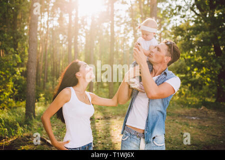 Elegante giovane famiglia mamma, papà e la figlia di un anno di età bionda seduta con il padre sulle spalle giocando felice e sorridente, all'esterno al di fuori della città Foto Stock
