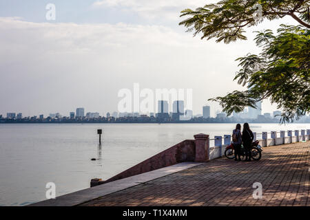 Due ragazze e una moto vicino Quan Tay Ho oppure Westlake del distretto di Hanoi. In riva al lago dei Tay con edifici residenziali e ristoranti. Il Vietnam. Foto Stock