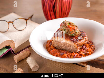Pancetta di maiale con lo stufato di fagioli e purea di verdure, servita in una piastra bianca, decorata con libro e bicchieri su un tavolo di legno. Foto Stock