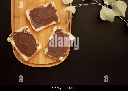 Una composizione di tre toast bianchi cosparsi di burro al cioccolato che giacciono su un tagliere contro con foglie uno sfondo scuro. vista superiore con sono Foto Stock