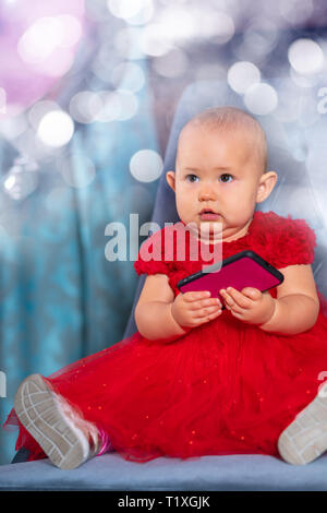 Little Baby girl in un colorato abito rosso festeggia il suo primo compleanno seduto in una sedia con party palloncini e frizzante bokeh di fondo Foto Stock