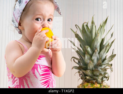 Piccolo Grazioso baby girl eating sour limone, tenendolo con entrambe le mani. Foglie di ananas maturo è in primo piano Foto Stock