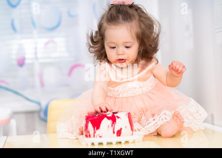 Confuso graziosa bambina in una rosa colore tonaca di mangiare una torta di compleanno con marmellata cosparsa attorno alla sua bocca e mani appiccicose come si siede lungo di esso su un tavolo Foto Stock