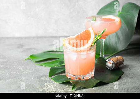 Rinfrescanti cocktail estivi di succo di pompelmo e ghiaccio calici in vetro grigio di un sottofondo di calcestruzzo con foglie di monstera. Foto Stock