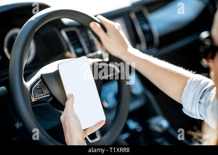 Donna che mantiene un telefono intelligente con schermata vuota a copia incolla durante la guida di una vettura sul volante Foto Stock