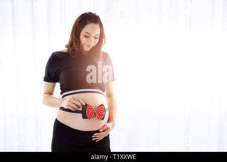 Giovane donna incinta mentre tiene il suo pancino con un arco di nastro al di sopra della finestra vista in mattinata con spazio di copia Foto Stock