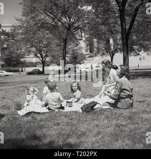 Degli anni Cinquanta, estate e due madri con i loro bambini seduti su un tappeto avente un picnic nel parco di fronte al Campidoglio di Washington DC, USA, la casa del governo statunitense. Foto Stock