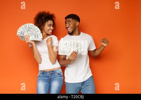 Grande fortuna. Allegro afro-americano di giovani l uomo e la donna godendo di un sacco di soldi, sfondo arancione Foto Stock