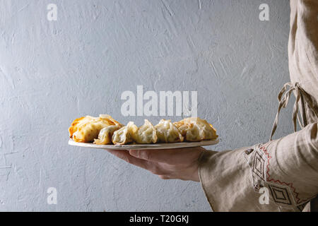 Fritti gnocchi asiatici Gyozas potstickers su bianco Vassoio in ceramica in mani femminili. Muro grigio a sfondo. Cena asiatica Foto Stock
