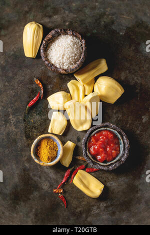 Gli ingredienti per cucinare vegan curry. Raw maturo pelato jackfruit con il bianco riso crudo, tagliate a pezzetti i pomodori e spezie in ciotole di ceramica su metallo scuro Foto Stock