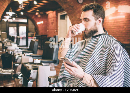 Uomo attraente è seduto nella sedia di barbiere e bere un tè. Anche lui sta guardando il telefono e leggere qualcosa da essa. Egli è in attesa f Foto Stock