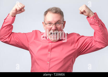 Felice senior bel Uomo in camicia rossa e bicchieri gesticolando e mantenendo la bocca. Egli è felice e celebra il suo successo. Foto Stock