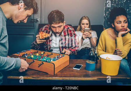 Due ragazzi stanno giocando a bordo del gioco del calcio. Essi sono molto concentrata su di esso mentre le ragazze stanno a guardare nel telefono e sento noioso. A loro non piace questo Foto Stock