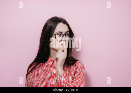 Close up ritratto di giovane e bella donna nel round di eyewear, tenendo il suo dito indice sulle labbra di mantenere il silenzio, vestito in camicia, ponendo su rosa Foto Stock