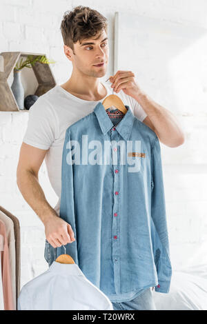 Uomo bello in t-shirt bianco azienda e cercando su camicie in camera da letto Foto Stock