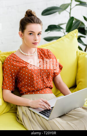 Fiducioso di moda giovane donna nella collana di perle seduto sul divano giallo e utilizzo di computer portatile Foto Stock