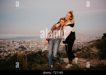 Sud Africa, Cape Town, Kloof Nek, due felici donne abbracciando al tramonto con il paesaggio urbano in background Foto Stock
