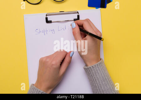 Donna mani di scrivere un elenco di acquisti su sfondo giallo. Spazio libero. Copia dello spazio. Flatlay. Foto Stock