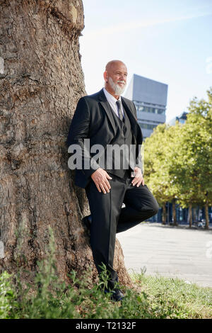 Imprenditore elegante poggiando su albero in città, pensando Foto Stock