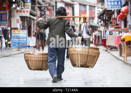 Uomo con polo sulla spalla con due cestelli appesi porta oggetti Market street in un tradizionale vecchio modo Foto Stock