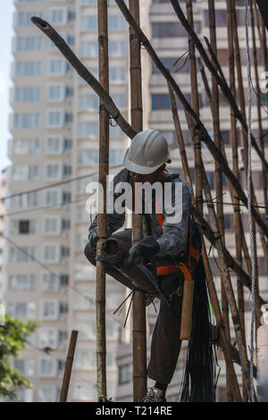 Un operaio edile che indossa un elmetto e un'imbracatura di sicurezza fissa ponteggi in bambù con lacci nere, garantendo la stabilità della struttura per i lavori di costruzione in corso. Foto Stock