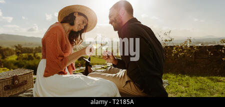 Giovane seduto su erba in un vigneto di vino di tostatura. Donna sorridente in hat seduto con il suo ragazzo beve il vino e a parlare con lui.