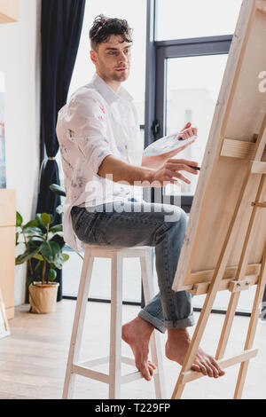 Bello artista in una camicia bianca e jeans blu dipinto su tela in galleria Foto Stock