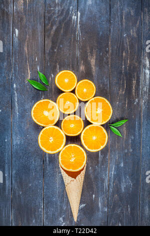 Creative stile scuro flat top laici vista di arance fresche fette di frutta nel gelato cono di cialda marrone sul tavolo di legno sfondo con copia spazio. Il minimo Foto Stock