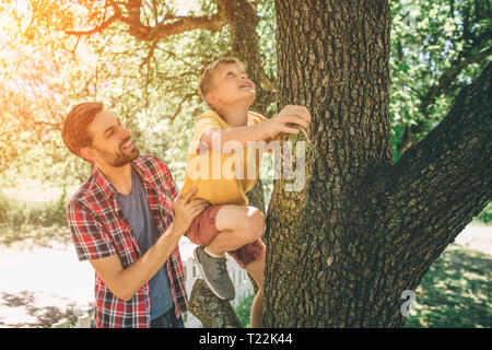 Padre Felice è aiutare il suo figlio a salire verso la cima dell'albero. Intrepido boy è in salita. Non ha paura. Ragazzo è guardando verso l'alto. Stanno giocando al di fuori in posizione di parcheggio Foto Stock