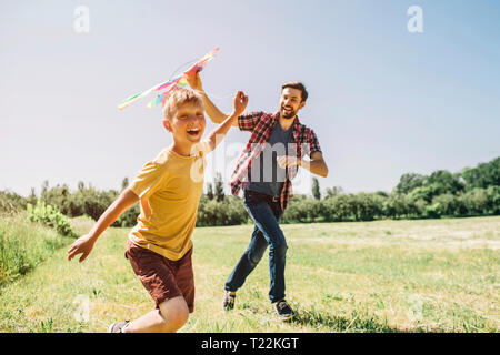 Capretto felice è in esecuzione sul campo. Egli è in possesso di thread da kite. Padre sta tenendo il kite. Egli sta cercando di eseguirlo al cielo. Essi stanno godendo il momento Foto Stock