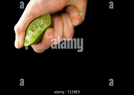 La mano di un uomo spreme il succo dalla metà di un lime. Succosa goccia a penzoloni. Isolare su sfondo nero. Foto Stock
