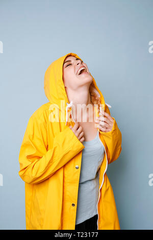 Ridendo giovane donna indossa giallo cappotto di pioggia di fronte a sfondo blu Foto Stock