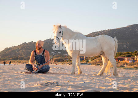 Spagna, Tarifa, uomo felice con pony seduto sulla spiaggia Foto Stock