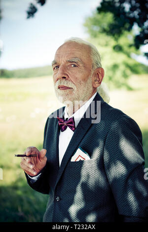 Ritratto di senior uomo indossare tuta coat, il filtro bow tie e abiti fazzoletto in natura fumatori sigaretto Foto Stock