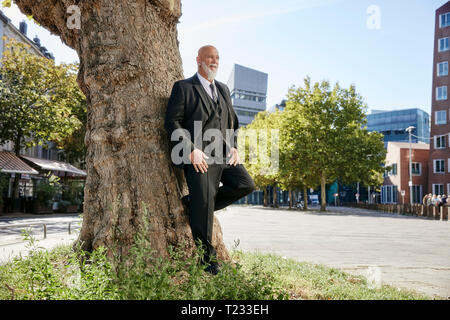 Imprenditore elegante poggiando su albero in città, pensando Foto Stock