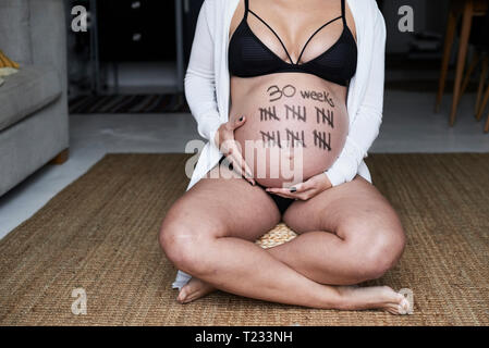 Close-up della donna incinta seduta sul pavimento a casa con i contrassegni di tally sul suo ventre Foto Stock
