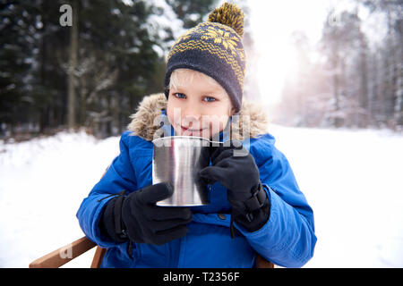 Ritratto di sorridere piccolo ragazzo seduto sulla slitta nella foresta di inverno bere il tè Foto Stock