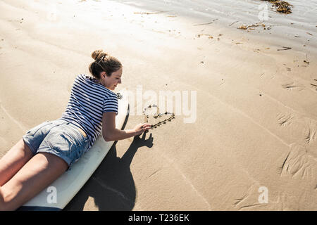 Giovane donna con la tavola da surf di disegno di un cuore nella sabbia Foto Stock