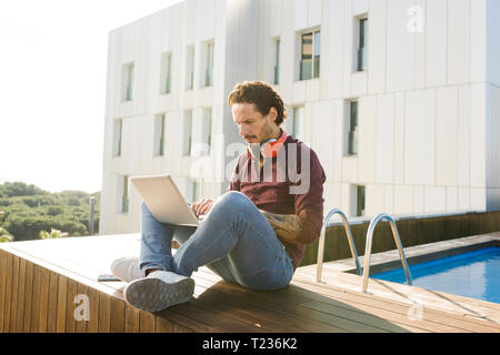 Uomo seduto a gambe incrociate su una terrazza sul tetto, utilizzando laptop, indossando le cuffie Foto Stock