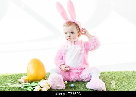 Carino baby in funny bunny costume seduta vicino le uova di Pasqua, tulipani e giallo uovo di struzzo isolato su bianco Foto Stock