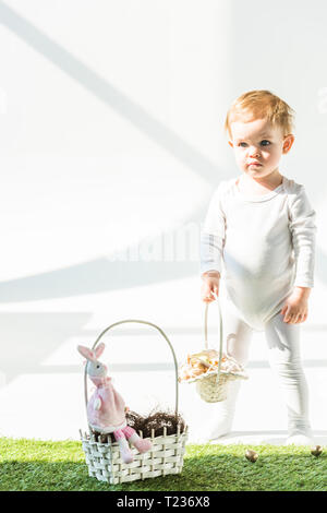 Carino baby azienda cesto di paglia mentre si sta in piedi sul prato verde in presenza di luce solare Foto Stock