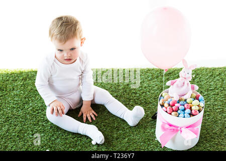Carino baby sitter su erba verde vicino a box con colorate uova di quaglia, coniglio decorativo e pallone aerostatico isolato su bianco Foto Stock