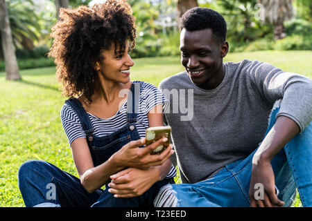 Felice coppia seduta sul prato in un parco, utilizza lo smartphone Foto Stock