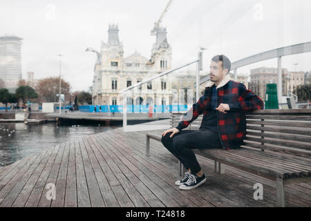 Giovane uomo che indossa abiti casual seduta su una panchina a porto Foto Stock