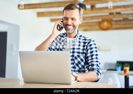 Uomo sorridente, parlando al telefono mentre si lavora da casa sul computer portatile Foto Stock